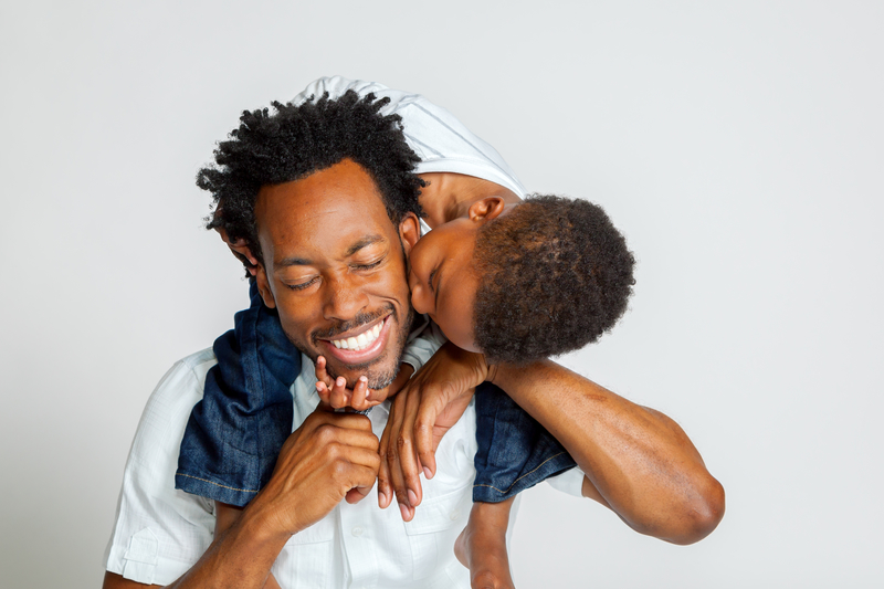 Young African-American adult with an African-American child on his shoulders. The boy is kissing his cheek, and he is smiling.