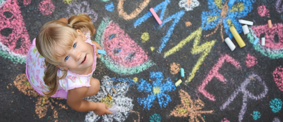 Young blonde girl holding blue chalk looks directly upward at the camera. She's standing on asphalt covered in chalk drawings of flowers, watermelons, and the word SUMMER.