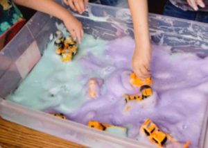 Children play with small cars and trucks in a plastic container filled with colored foam.