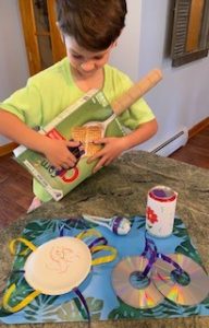 A young boy plays with a guitar he made at home with a cereal box and paper towel tube. He also shows instruments he made out of paper plates, tin cans, CDs, and plastic eggs.