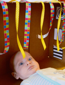 A baby, laying on his back, looks at brighly colored strips of ribbon attached to a box