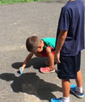 A young child traces another child's shadow with chalk on pavement