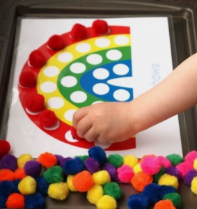 A child places red pom poms on the red stripe of a rainbow that is printed on a sheet of white paper