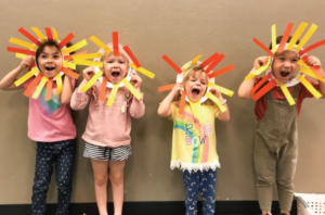 Four young children wearing lion masks made from yellow, orange, and white paper