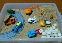 Toy cars and trucks in a clear plastic container filled with sand and rocks